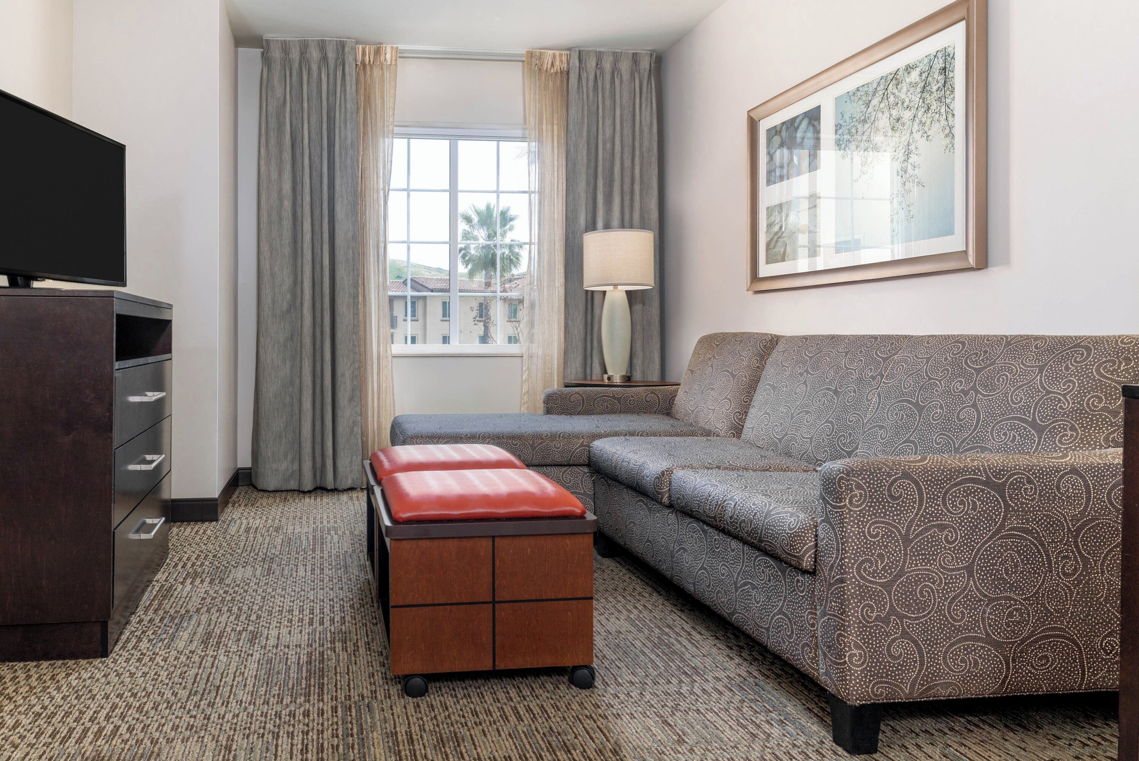 A hotel room with a sofa, a TV, a lamp, a small table with a red cushion, and a window with curtains.