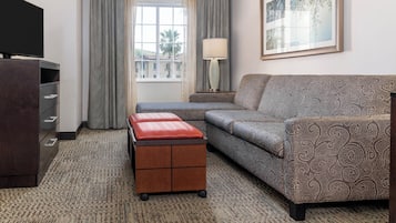 A hotel room with a sofa, a TV, a lamp, a small table with a red cushion, and a window with curtains.