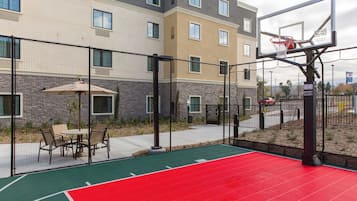 An outdoor basketball court with a red playing surface, surrounded by a fence, and adjacent to a building with multiple windows.