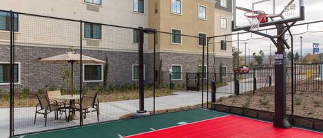 An outdoor basketball court with a red playing surface, surrounded by a fence, and adjacent to a building with multiple windows.