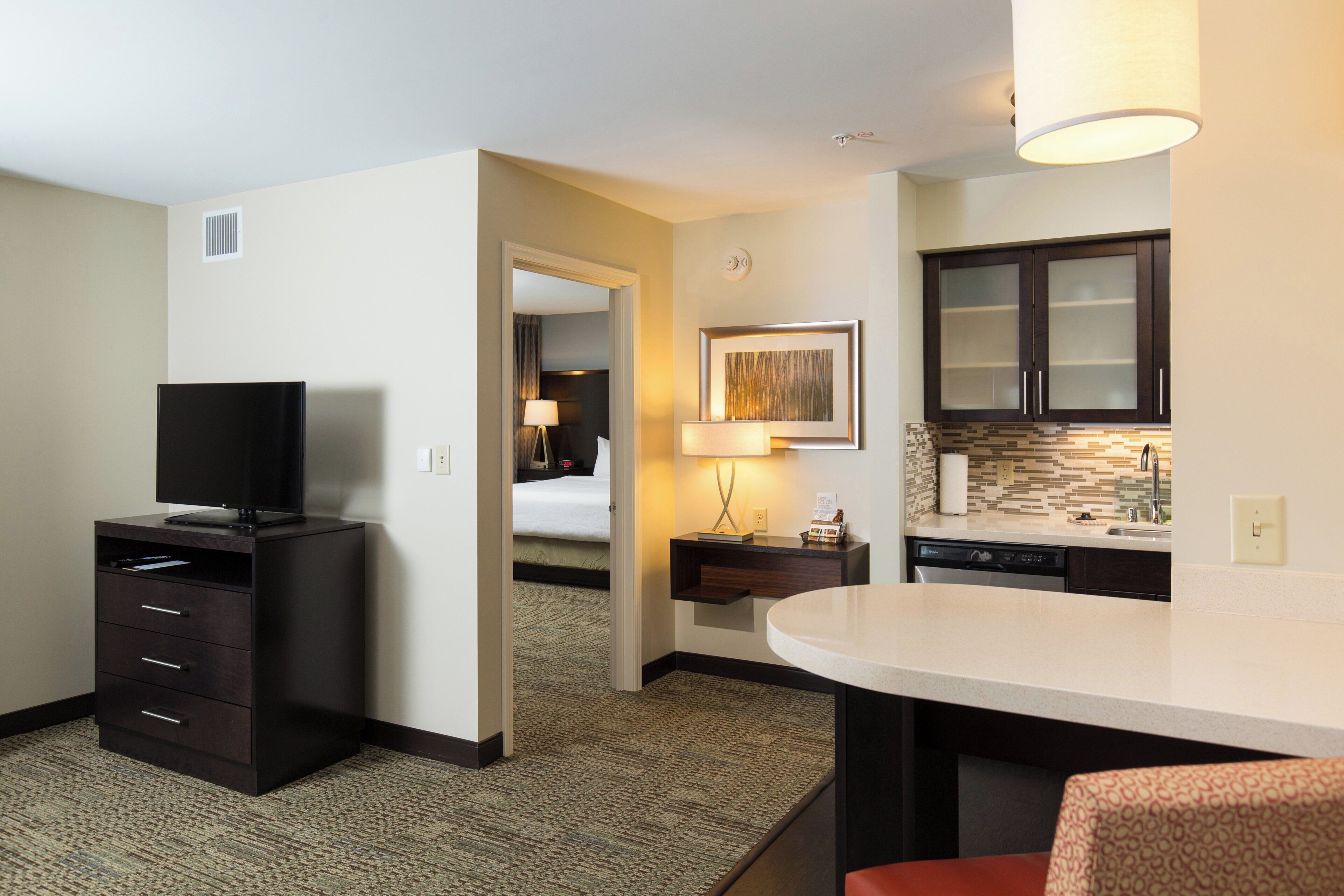 Habitación de hotel con televisión, escritorio con lámpara, cama y kitchenette.