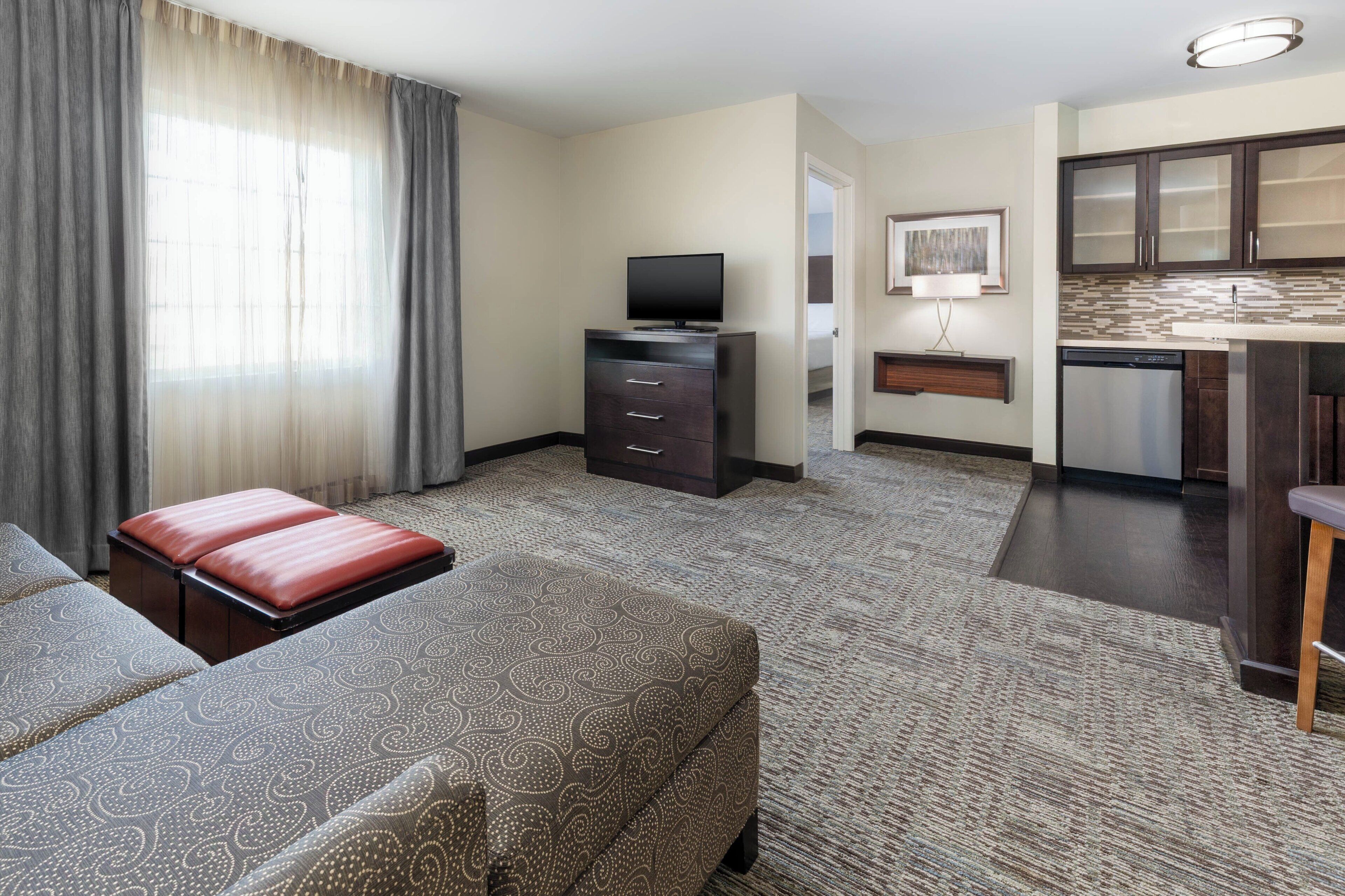 A hotel room with a sofa, a television, a kitchenette, and a large window with curtains.