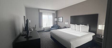 A hotel room with a large bed, a desk with a computer, a chair, a window with curtains, and a nightstand with a lamp.