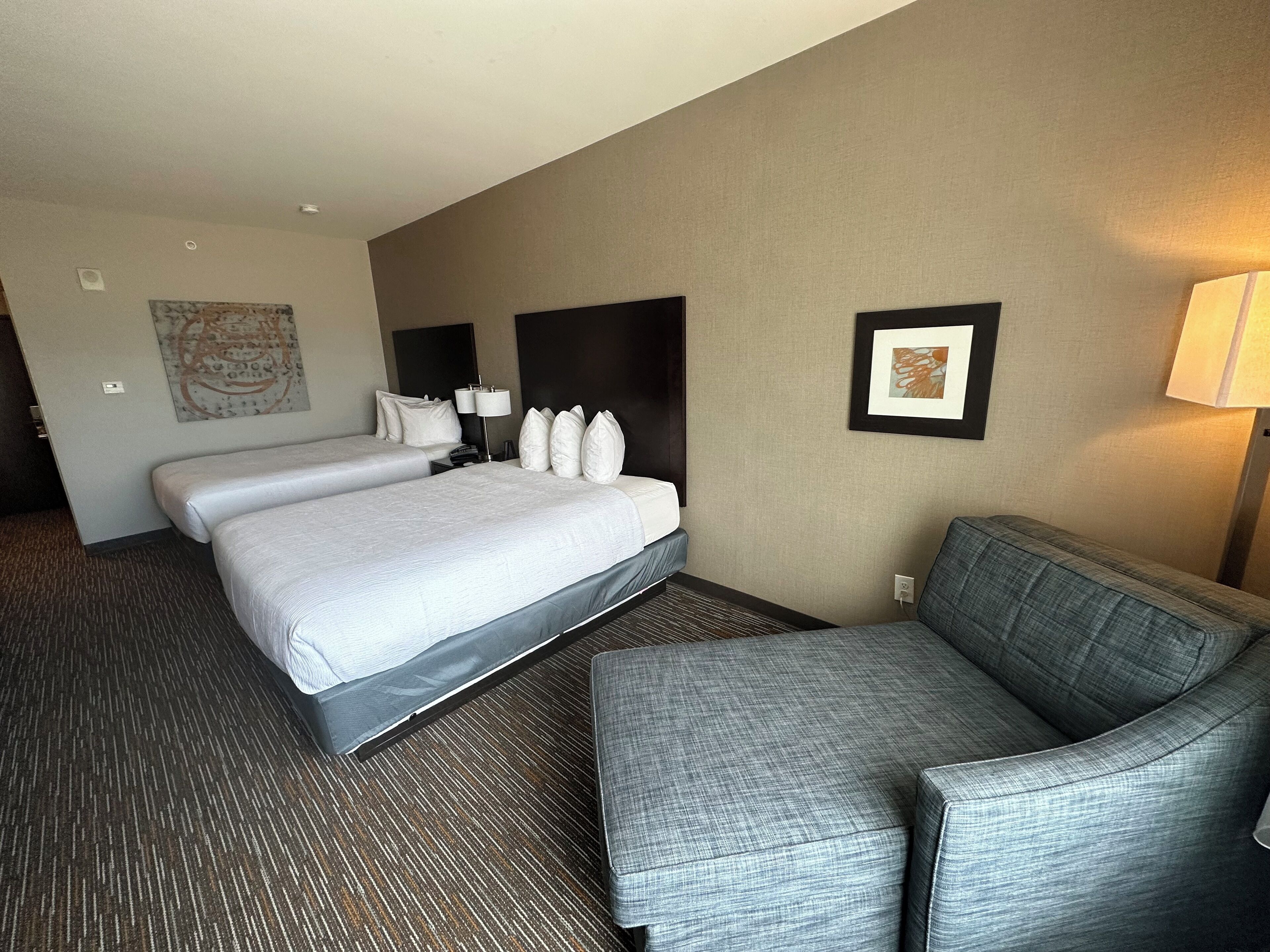 A hotel room with two beds, a gray armchair, a wall-mounted lamp, and a framed picture.