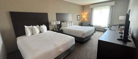 A hotel room with two beds, a desk with a computer, a lamp, and a window with curtains.