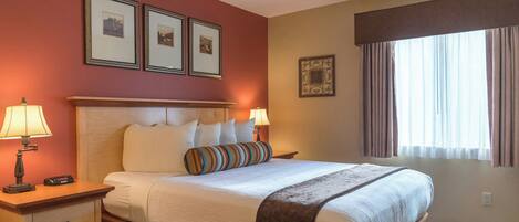 Habitación de hotel con cama, dos mesitas de noche, una lámpara, un control remoto, una ventana con cortinas y tres cuadros en la pared.