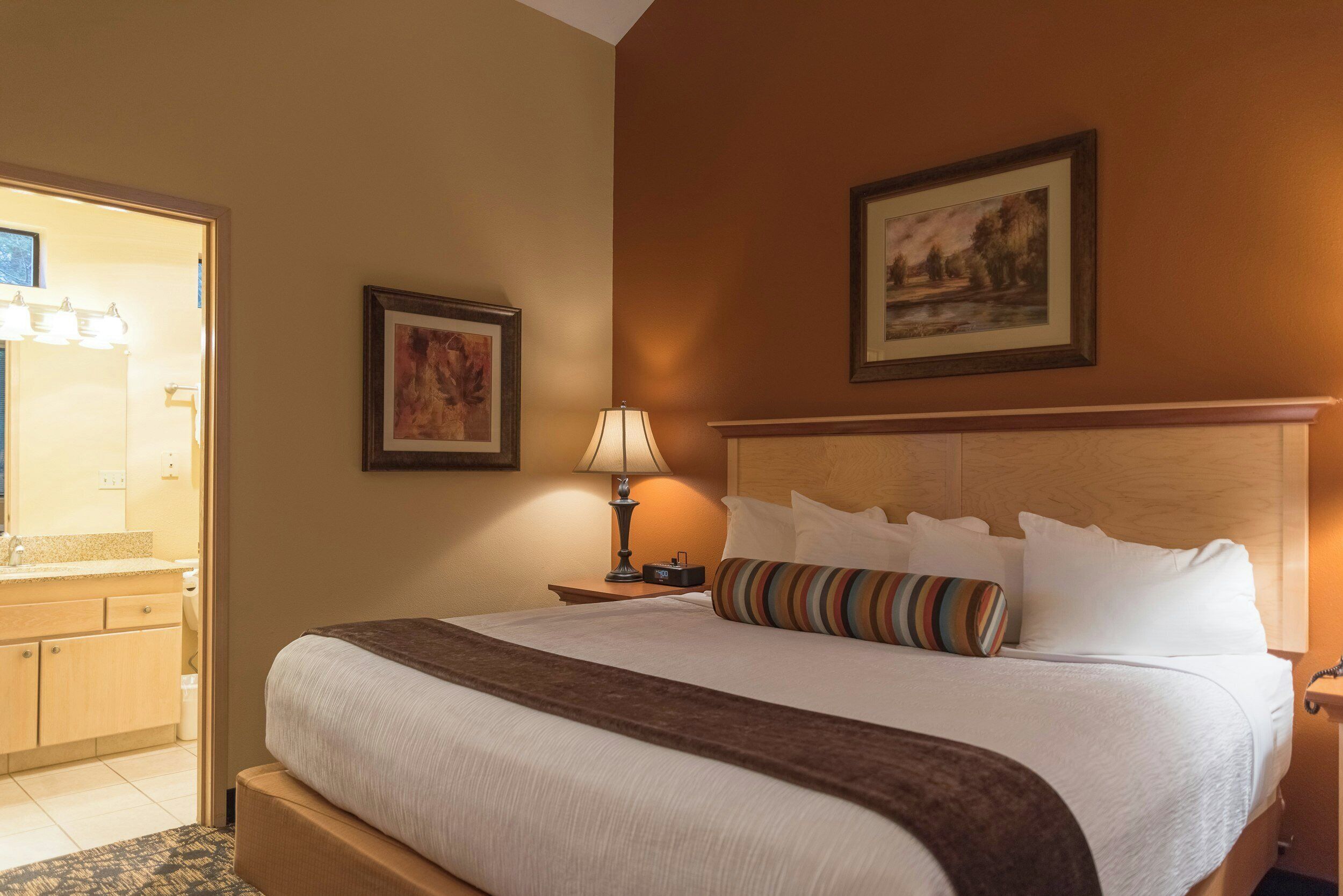 Hotelkamer met groot bed, nachtkastje met lamp, schilderij aan de muur en zichtbare badkamer door een open deur.