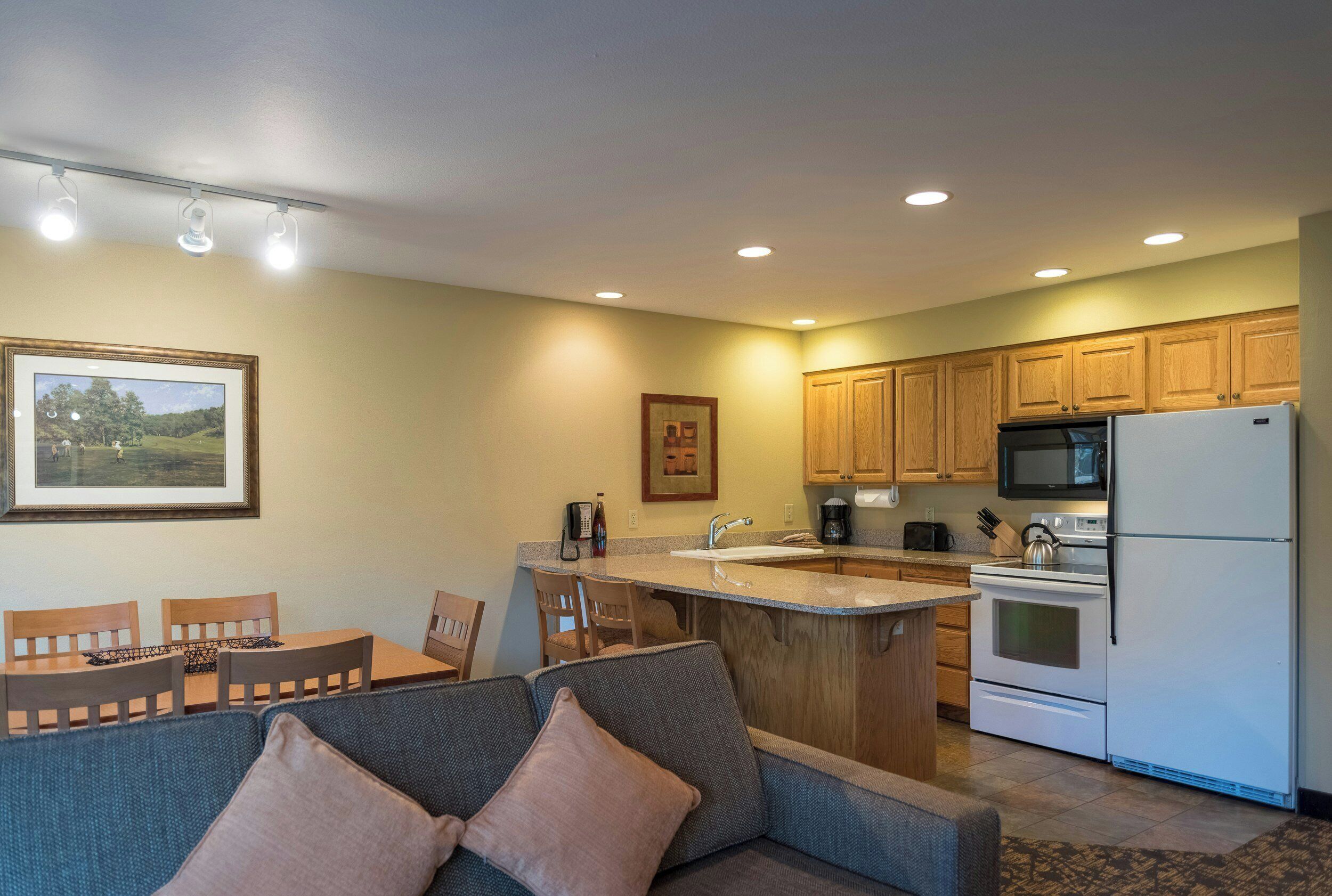 A compact kitchen with wooden cabinets, a built-in microwave, and a refrigerator. There is a dining area with a table and chairs, and a sofa with cushions.