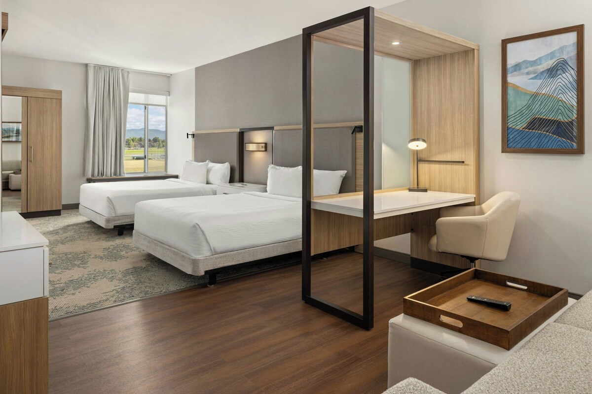 Habitación de hotel con dos camas, un escritorio, una silla, un espejo grande y vistas al exterior.