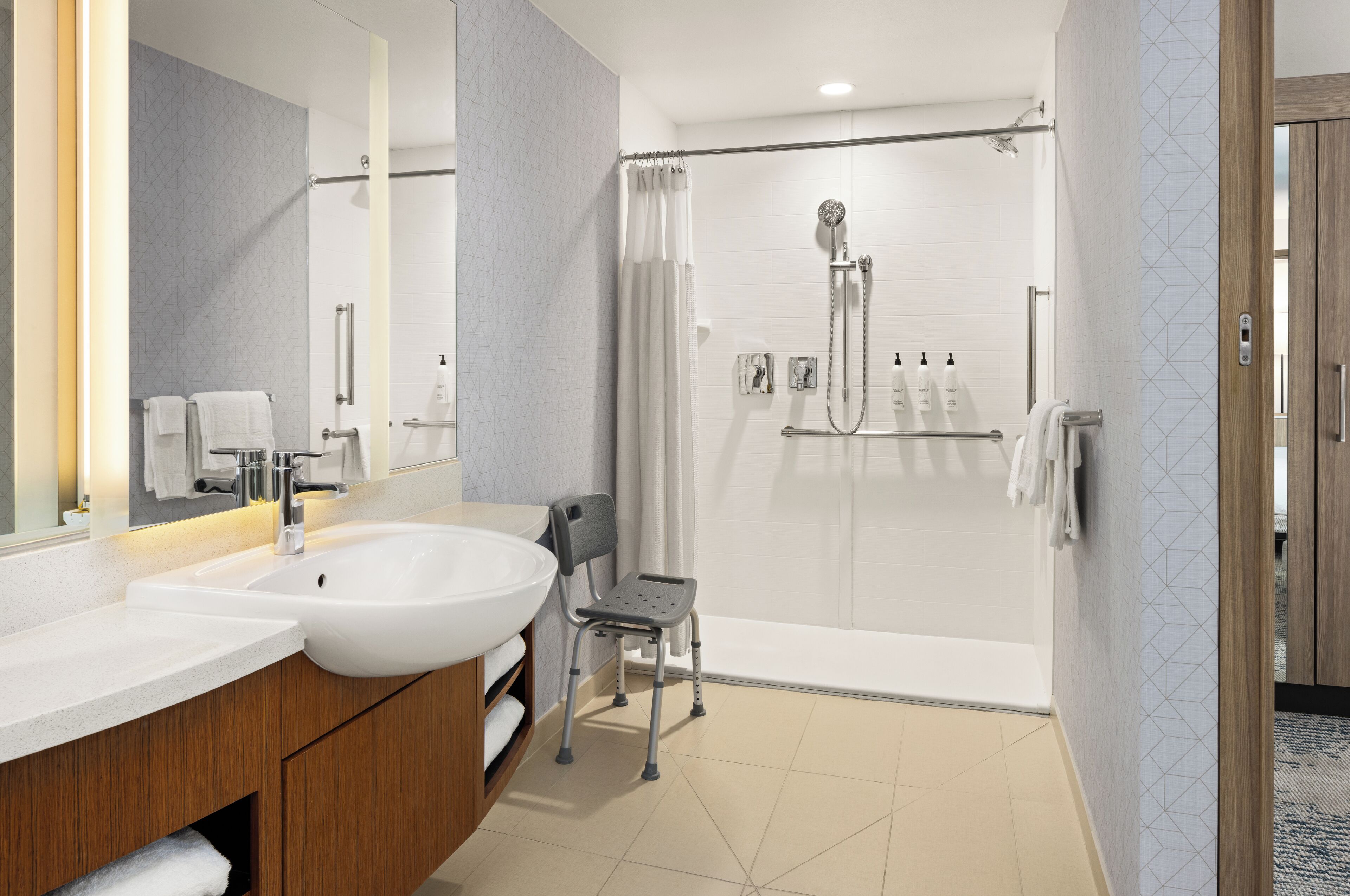 A modern bathroom with a large sink, mirror, shower, and a wheelchair-accessible chair.