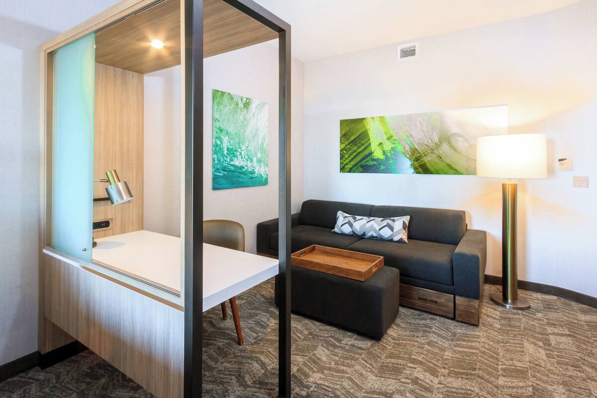 A modern hotel room with a wooden-framed bed, a white desk, a black sofa, and a floor lamp.