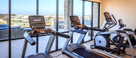 Ein Fitnessstudio mit Laufbändern, Crosstrainern und Rudergeräten. Große Fenster bieten einen Blick auf die Stadt.