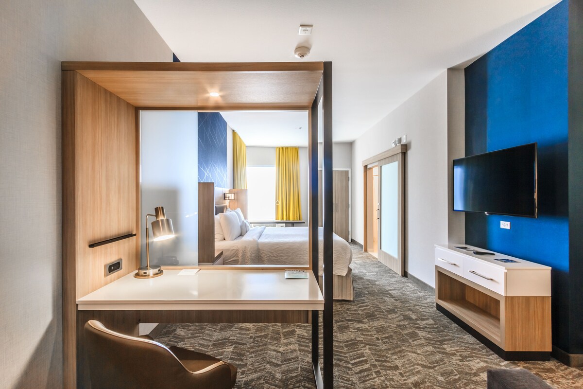 Une chambre d’hôtel équipée d’un bureau, d’une chaise, d’un lit, d’une télévision, et offrant une vue sur le couloir et une autre chambre.