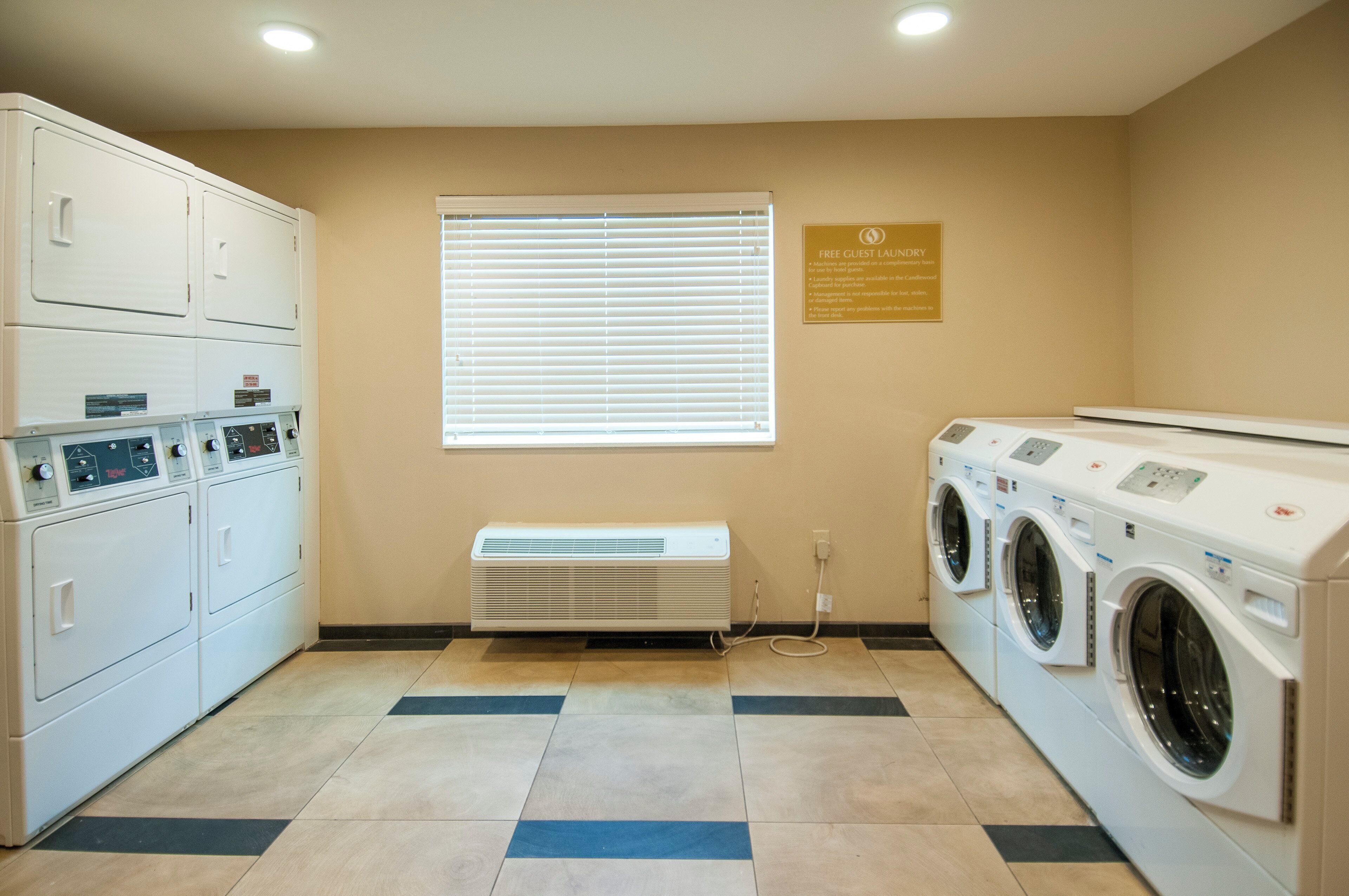 Un cuarto de lavandería con varias lavadoras y secadoras, una unidad de ventilación y un cartel que indica servicio de lavandería gratuito para huéspedes.