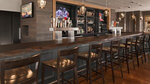 A bar area with a long counter, multiple bar stools, and a television screen displaying a sports event. - The Inn at Swarthmore (Swarthmore)