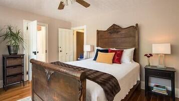 A bedroom with a wooden bed, white walls, a ceiling fan, and a nightstand with a lamp and books.