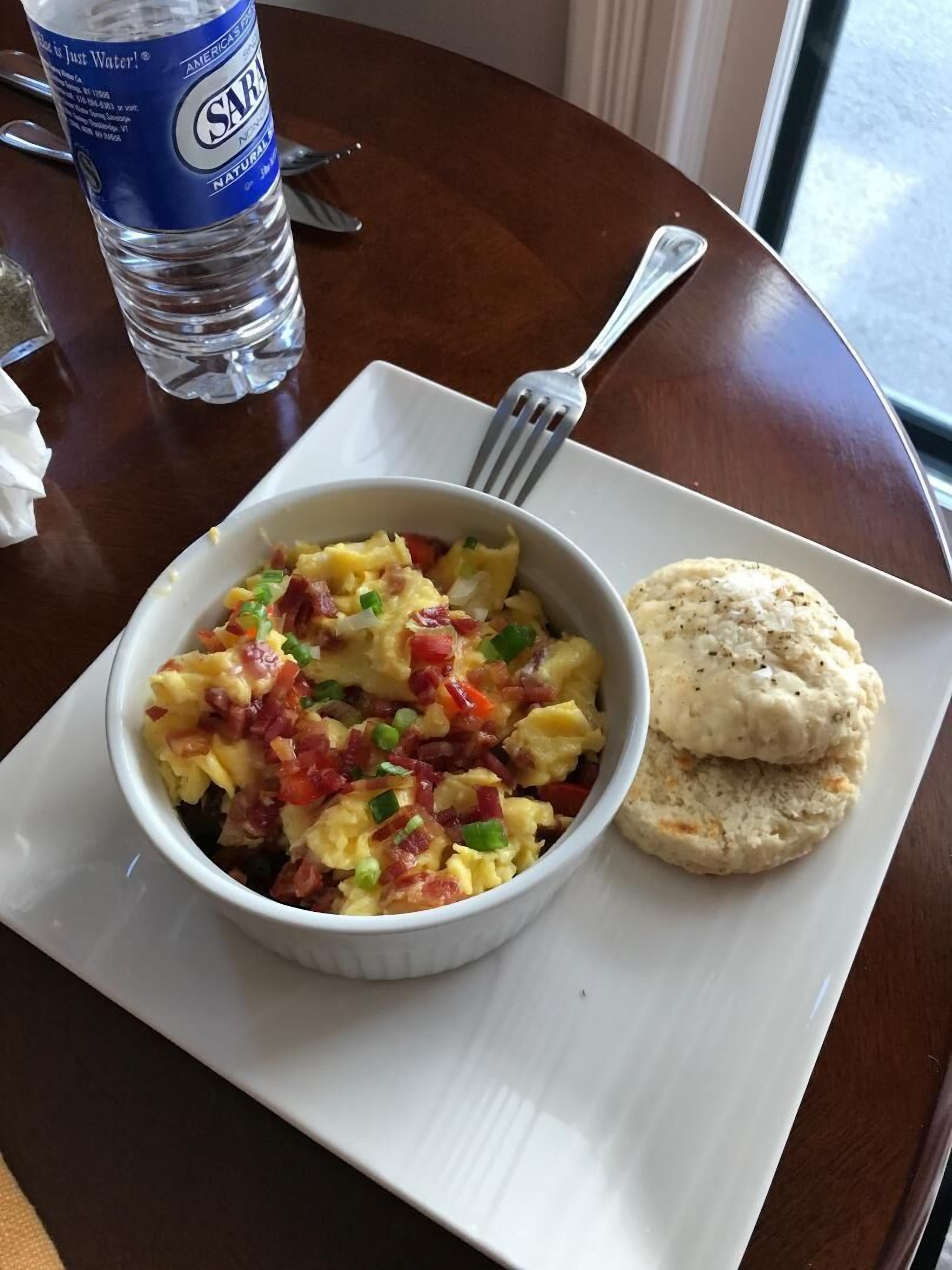 Une assiette de petit-déjeuner comprenant des œufs brouillés, du bacon et des poivrons verts, accompagnée d’un biscuit et d’une bouteille d’eau.