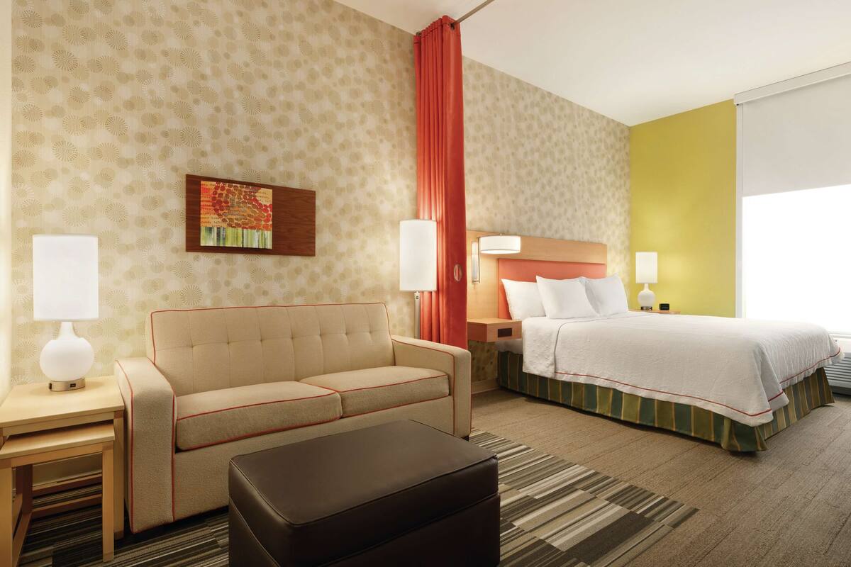 A hotel room with a sofa, a bed, a nightstand, a lamp, a painting, and a window with curtains.