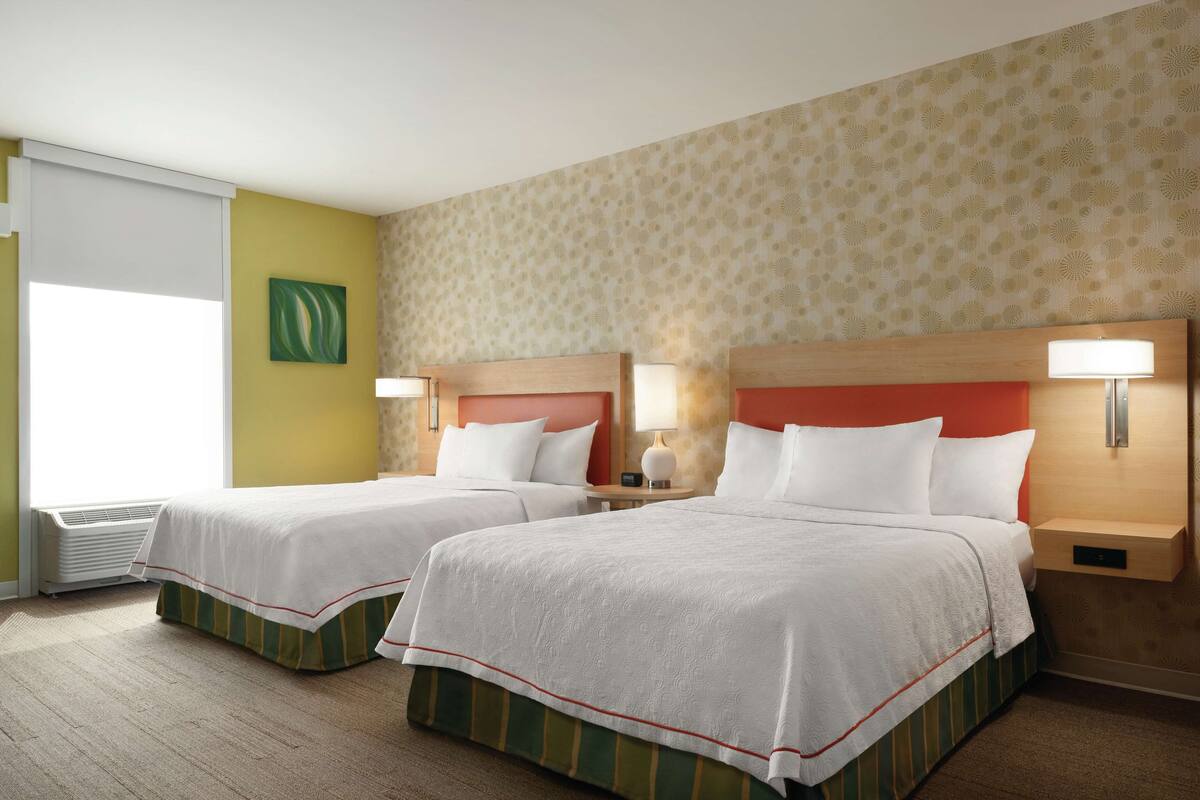 A hotel room with two beds, a window with blinds, a wall-mounted artwork, and a patterned wallpaper.