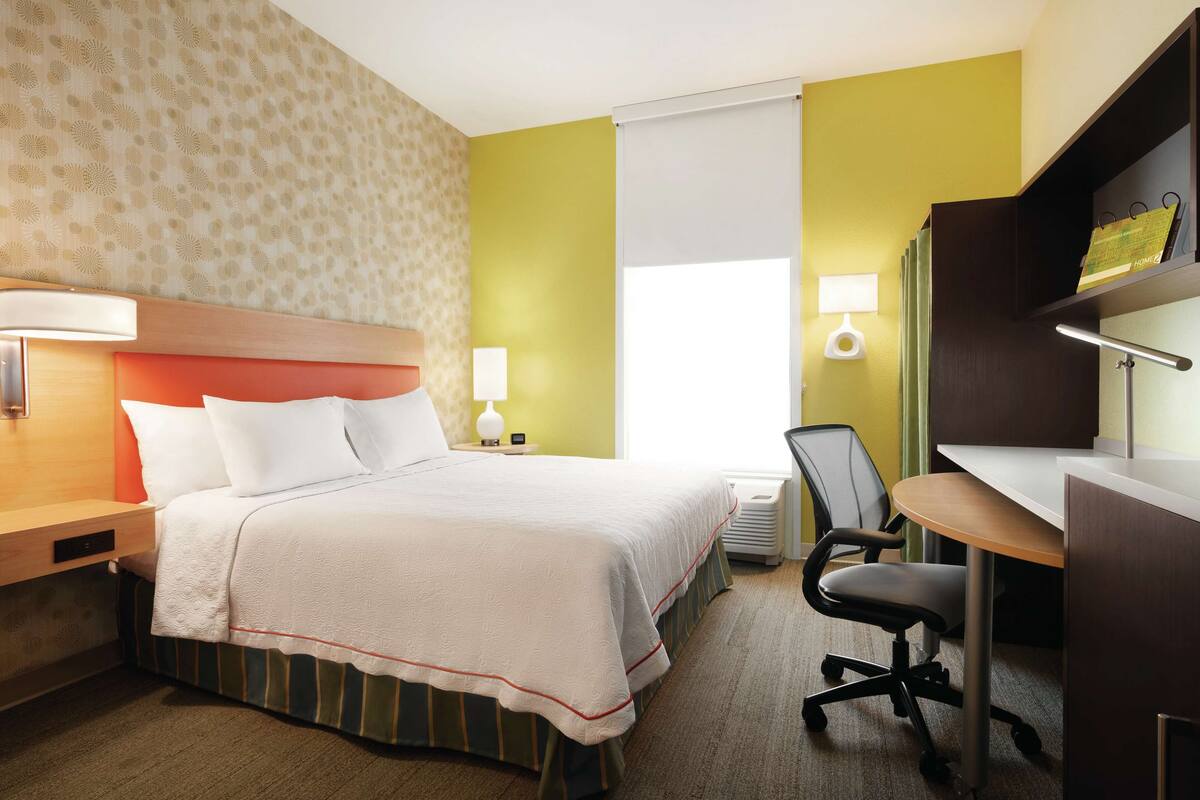 A hotel room with a bed, desk, chair, and a window with blinds.