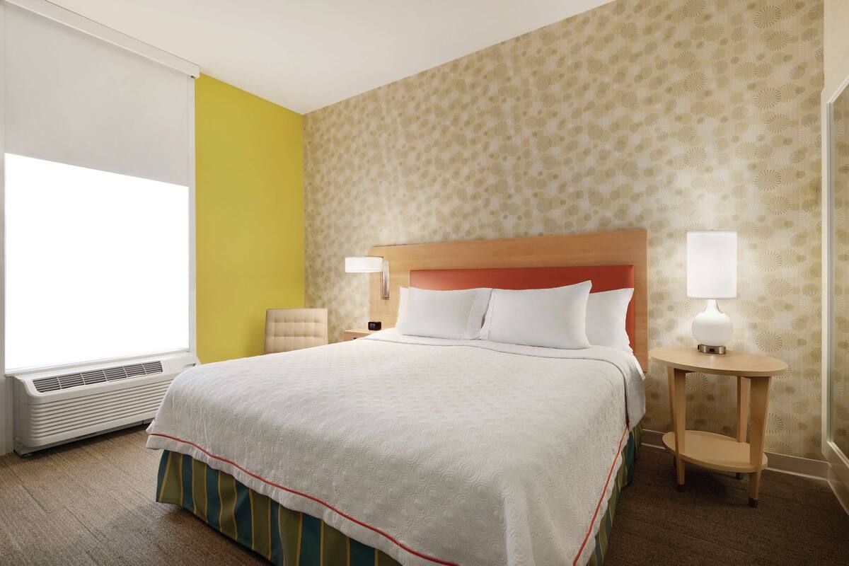A hotel room with a large bed, a bedside table with a lamp, a wall air conditioner, and a window with blinds.