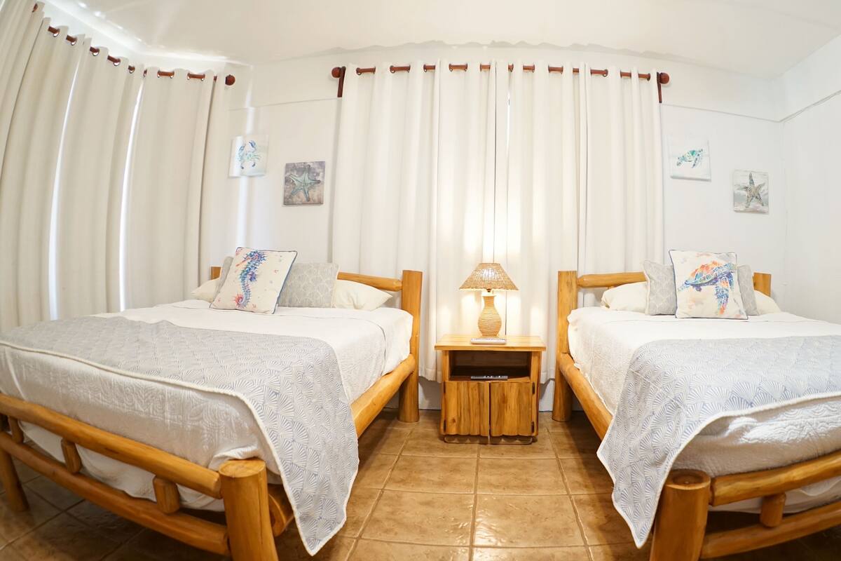 A double room with two wooden beds, a nightstand with a lamp, and a view of the sea through sheer curtains.