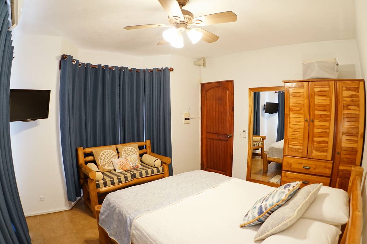 A bedroom with a bed, a wooden wardrobe, a ceiling fan, a chair, and a television on the wall.