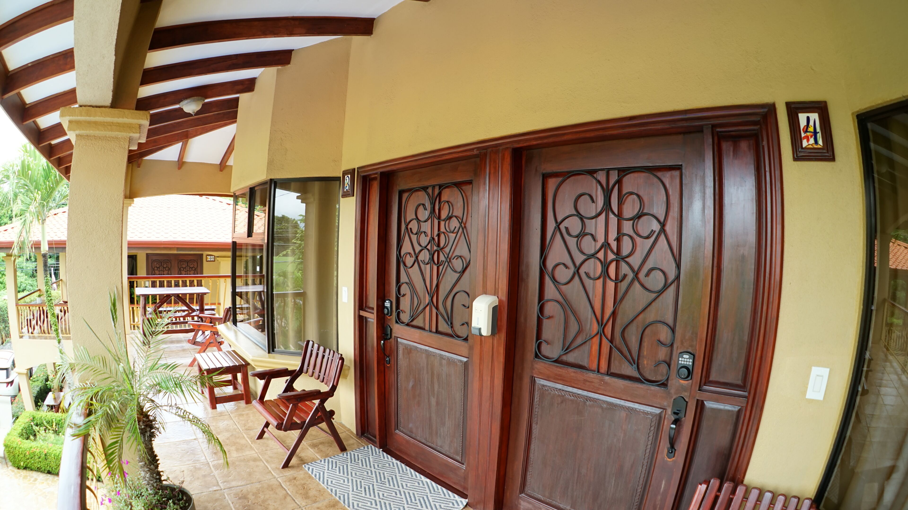 A wooden door with intricate metalwork, a bench, and a small table outside a building with a tiled floor and a covered patio area.