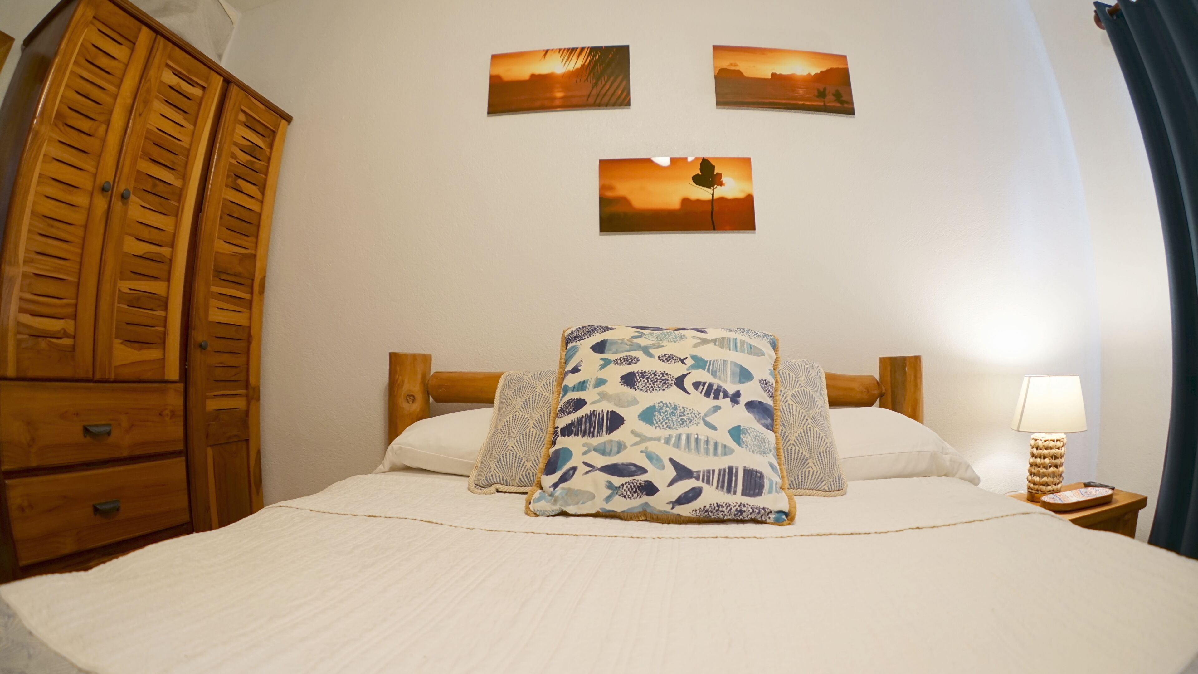 A bedroom with a bed, wooden wardrobe, and three wall-mounted pictures.