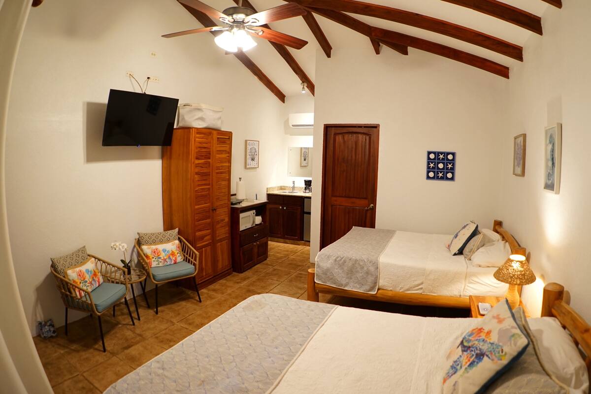 A bedroom with two beds, a wooden wardrobe, a chair, a desk with a computer, and a ceiling fan.