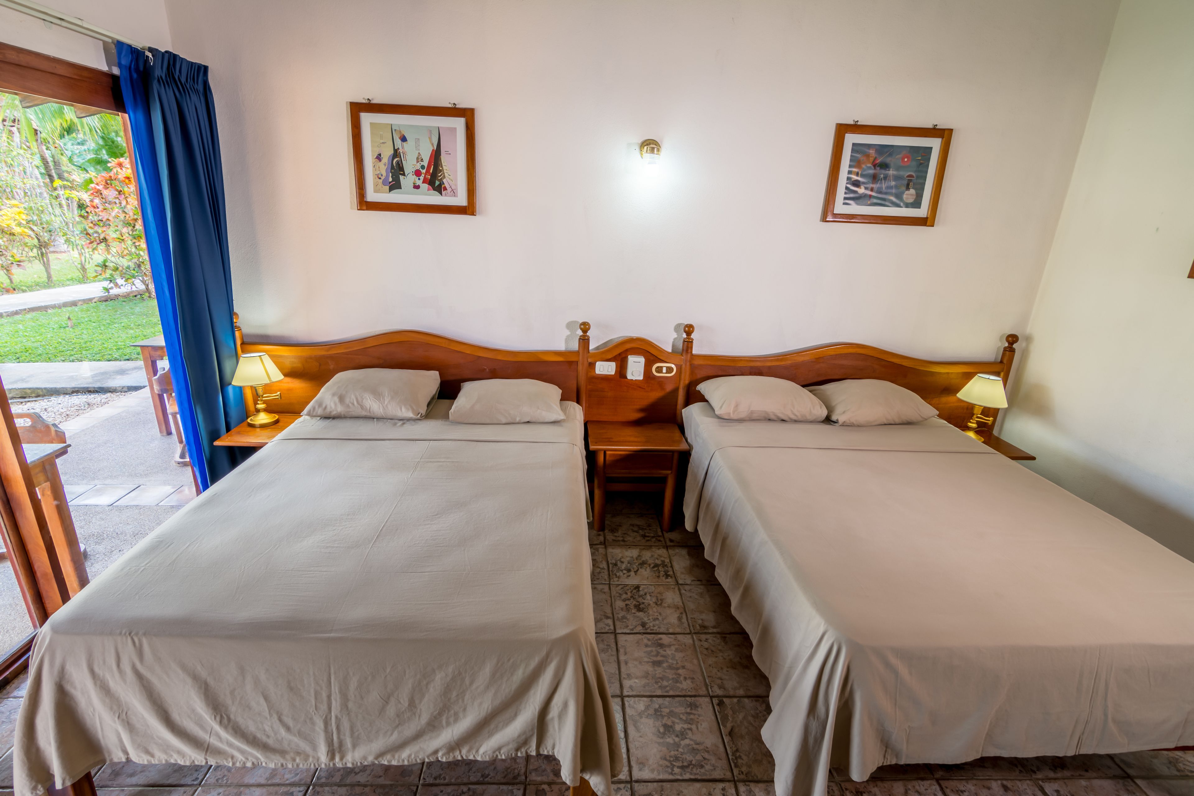 Une chambre d’hôtel avec deux lits simples, une tête de lit en bois, une table de chevet avec une lampe et une fenêtre avec un rideau bleu.