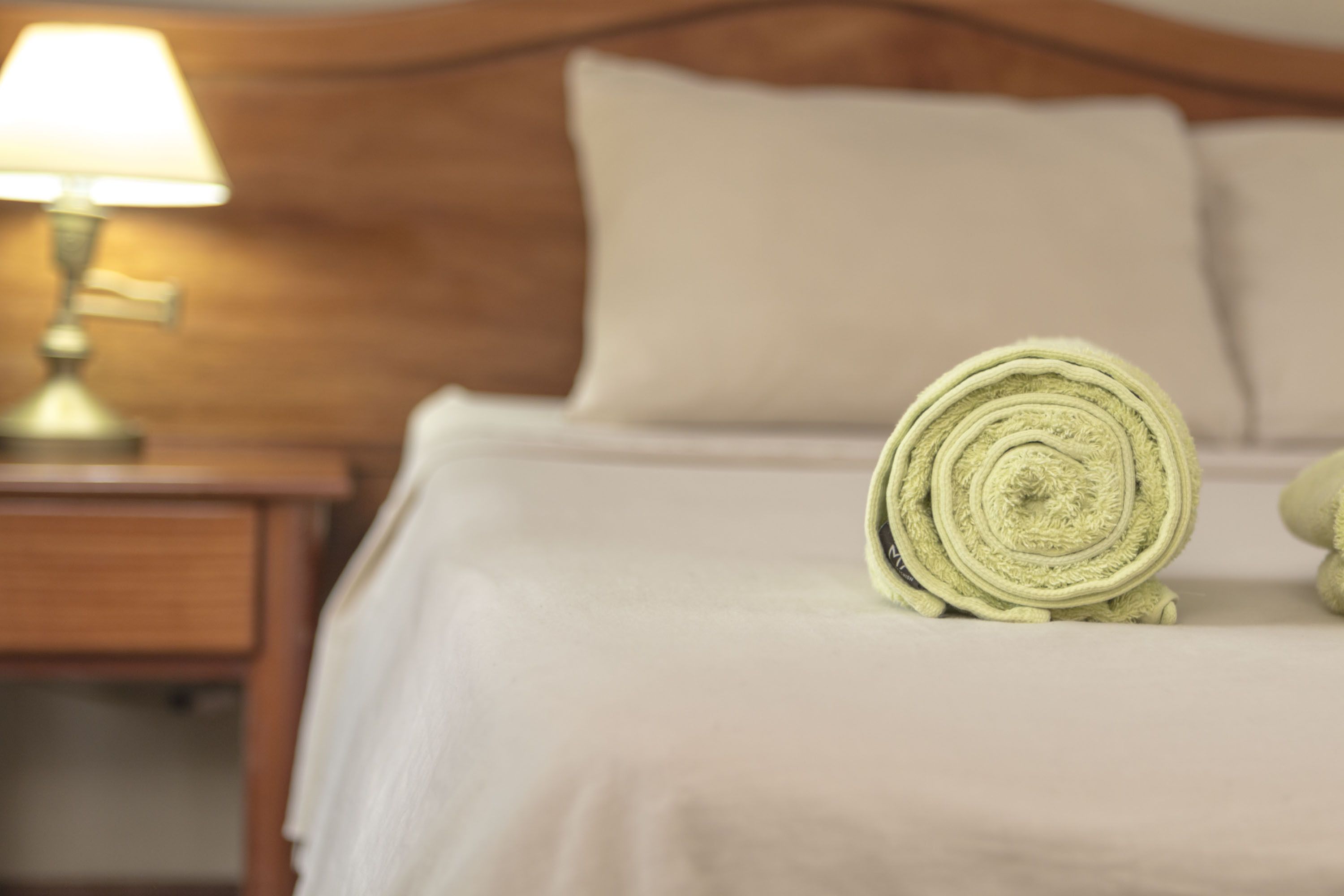 Een netjes opgemaakt bed met een opgerolde handdoek en een lamp op een houten nachtkastje.