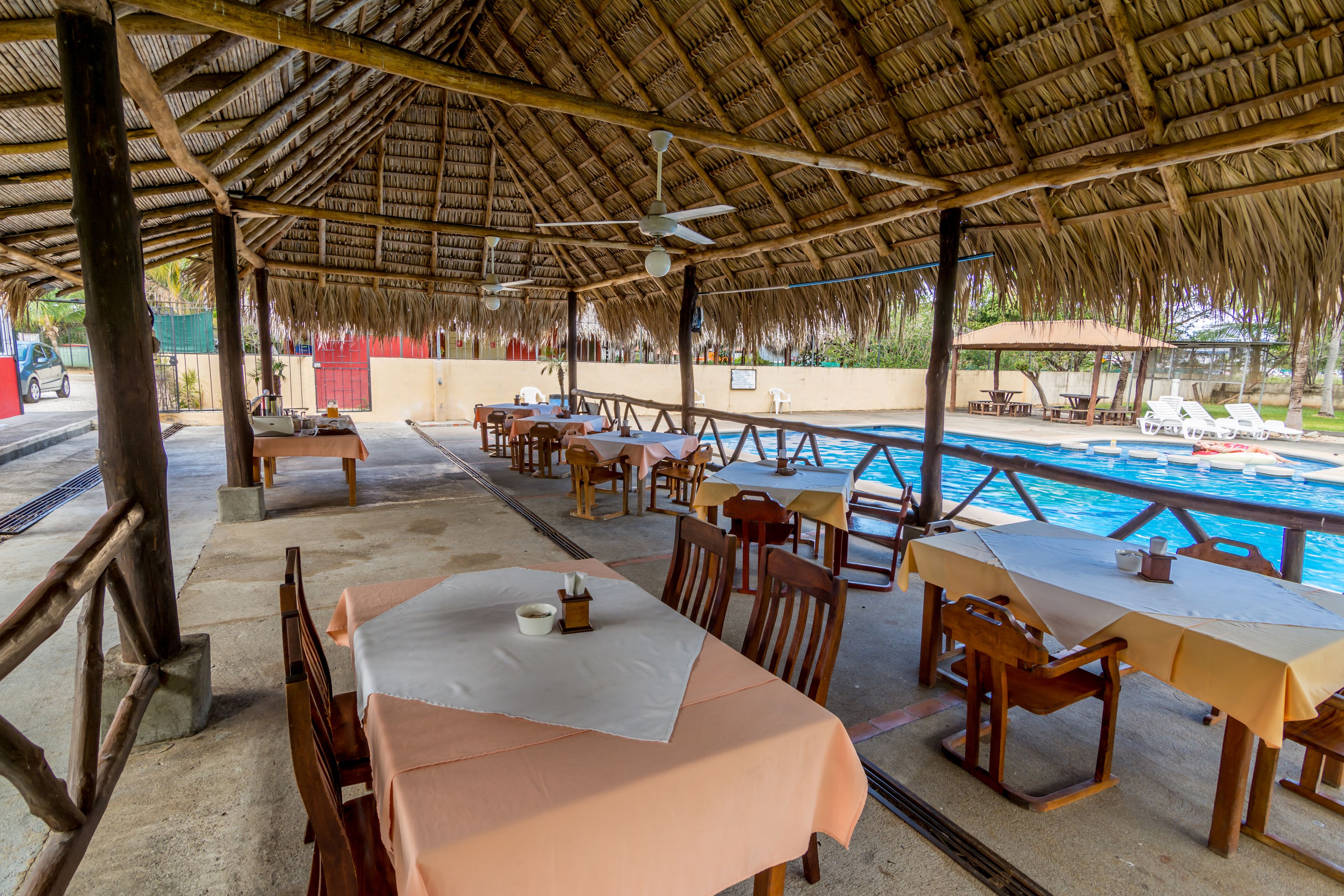 An outdoor dining area with tables and chairs, a pool in the background, and a thatched roof structure.