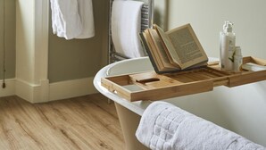 A bathroom with a wooden floor, a bathtub, a towel rack with two white robes, a shelf with a plant, and a towel rack with multiple towels. - Methuen Arms (Corsham)