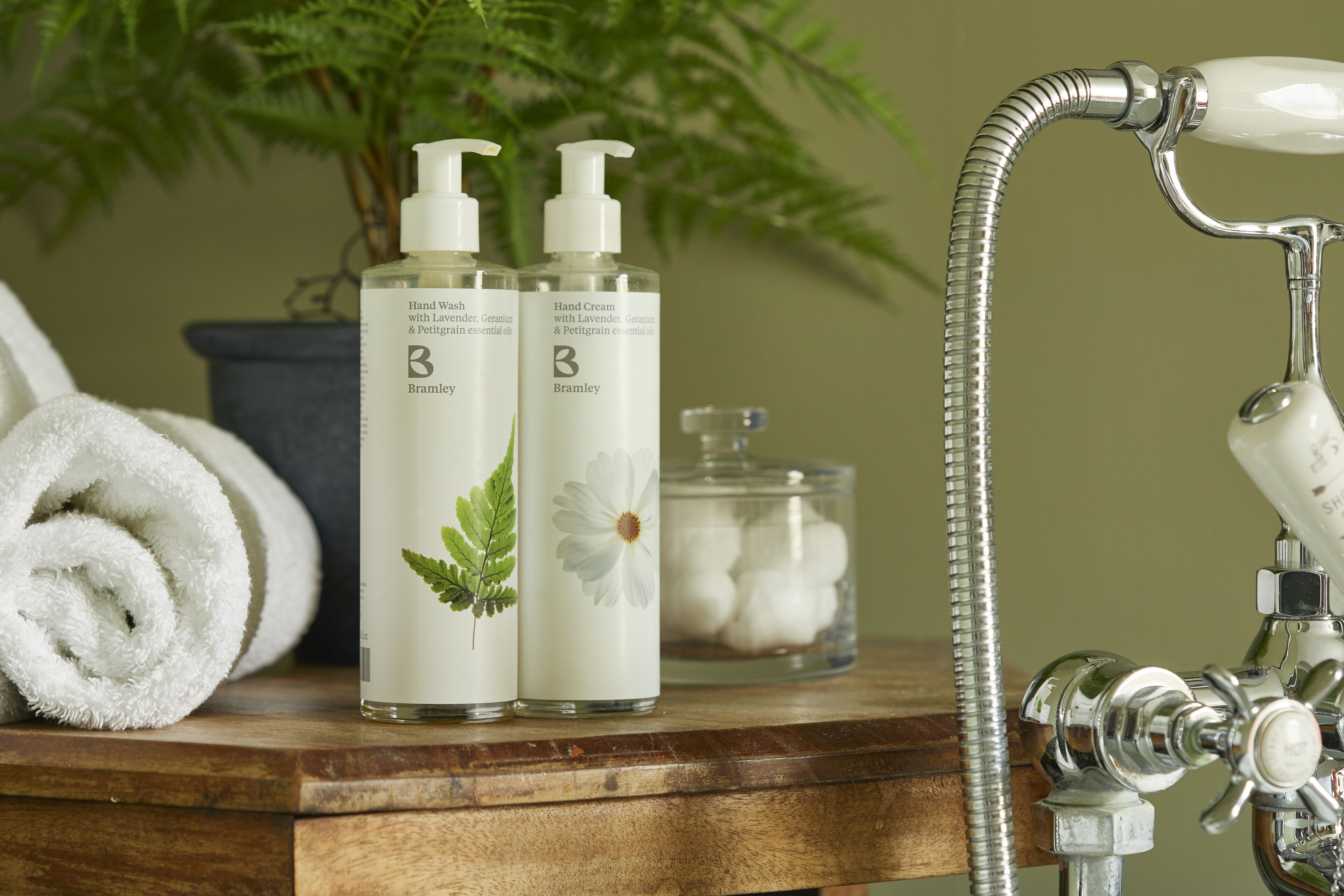 A bathroom vanity with two bottles of Bramley hand wash and hand cream, a rolled towel, a glass jar of cotton balls, and a showerhead.