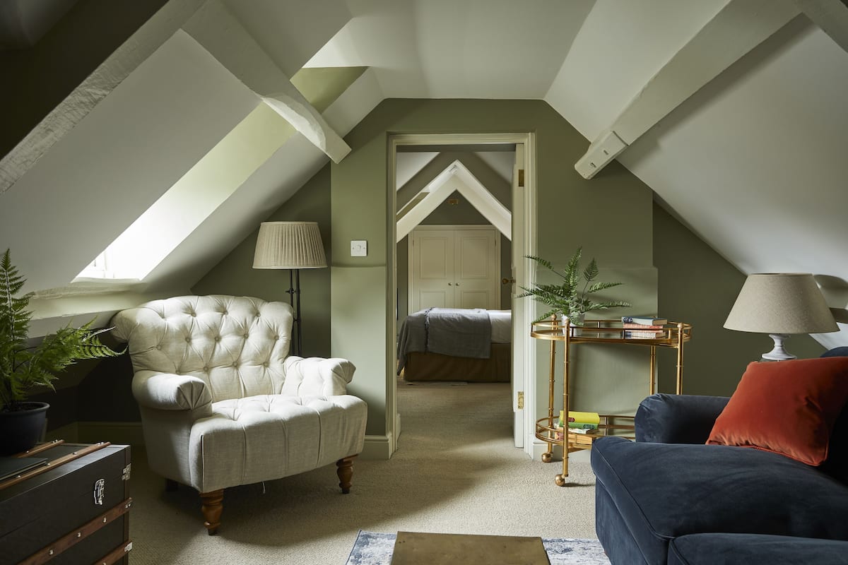 A loft-style room with a high ceiling, a tufted armchair, a sofa, a lamp, and a bookshelf.