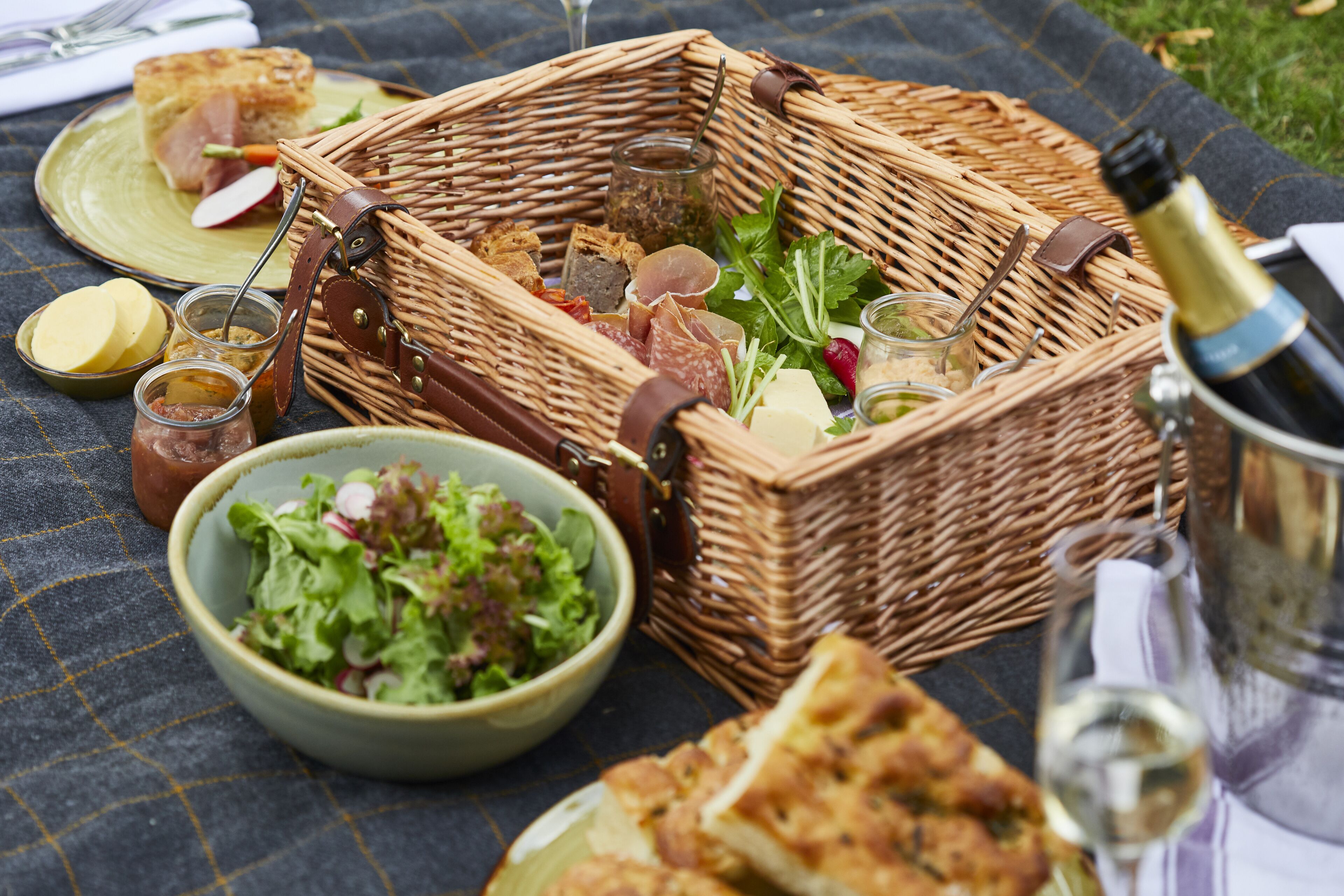 A picnic setup with a wicker basket containing food items, a bowl of salad, and a bottle of wine.