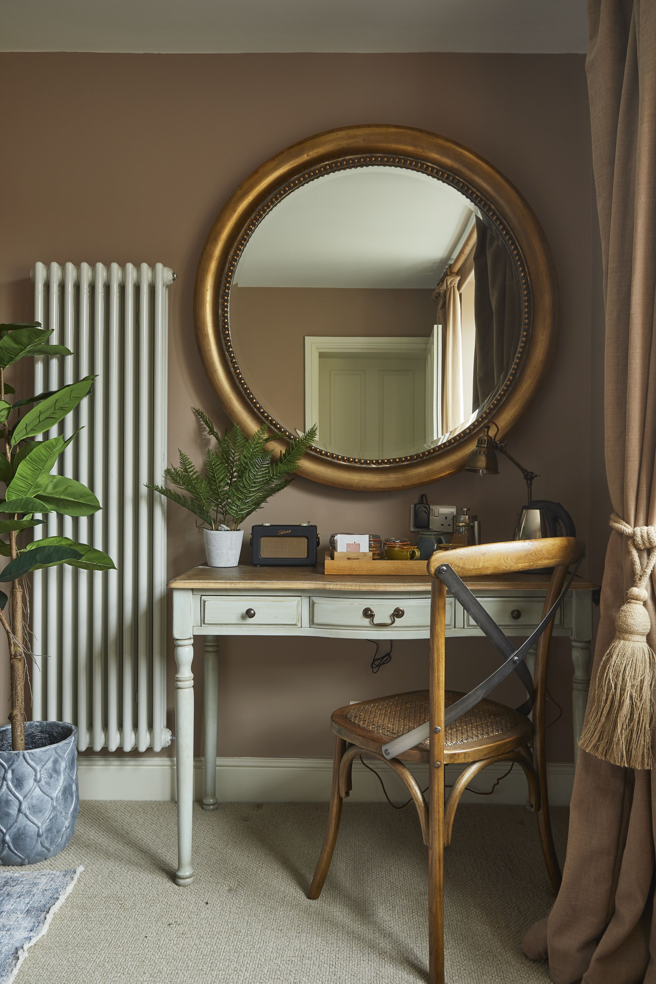 A room with a wooden chair, a white desk, a large mirror, a radiator, a potted plant, and a curtain.