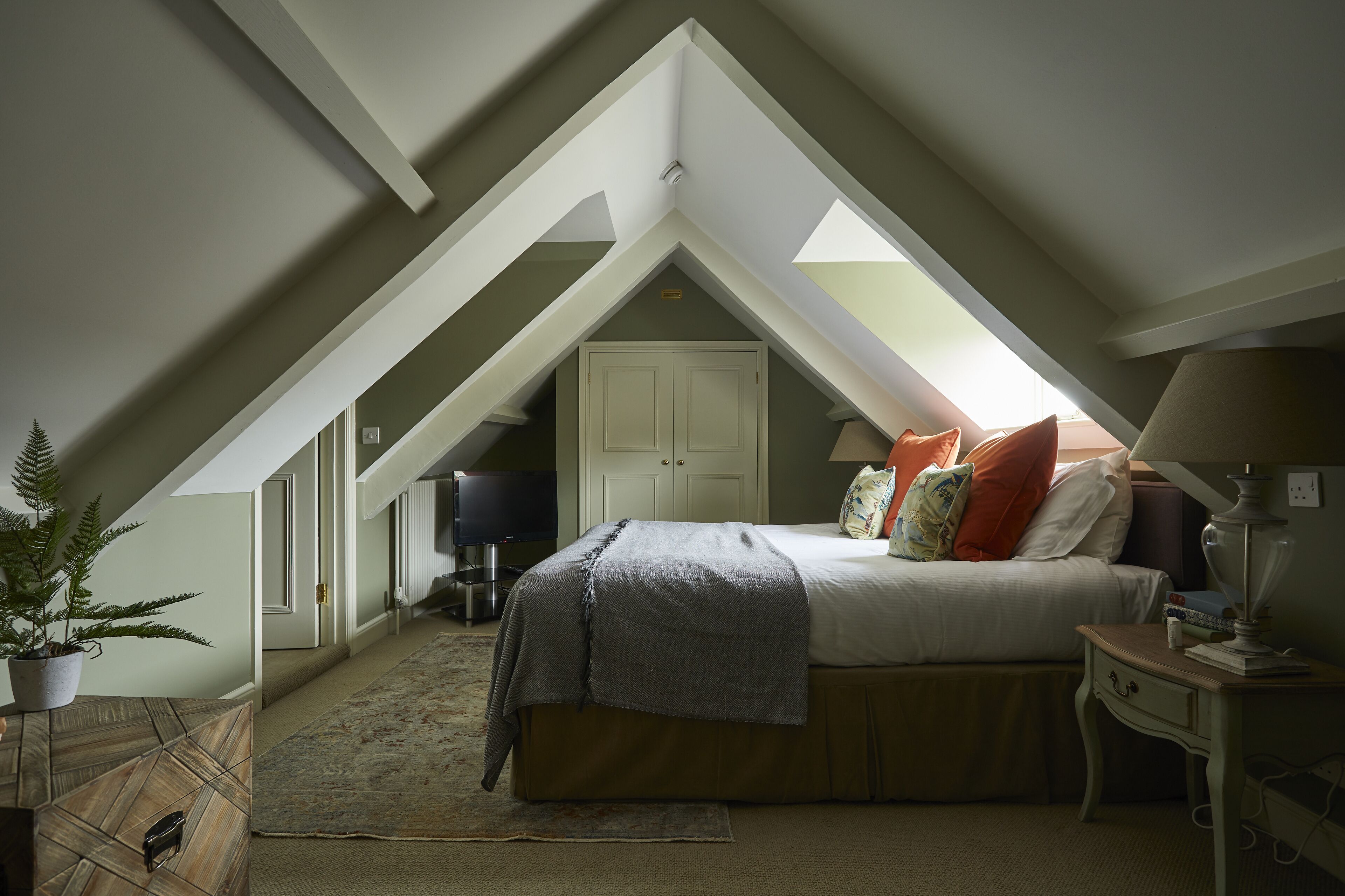 Ein Schlafzimmer mit schräg verlaufender Decke, einem Bett mit Kissen, einem Nachttisch, einer Lampe und einem Blumentopf.