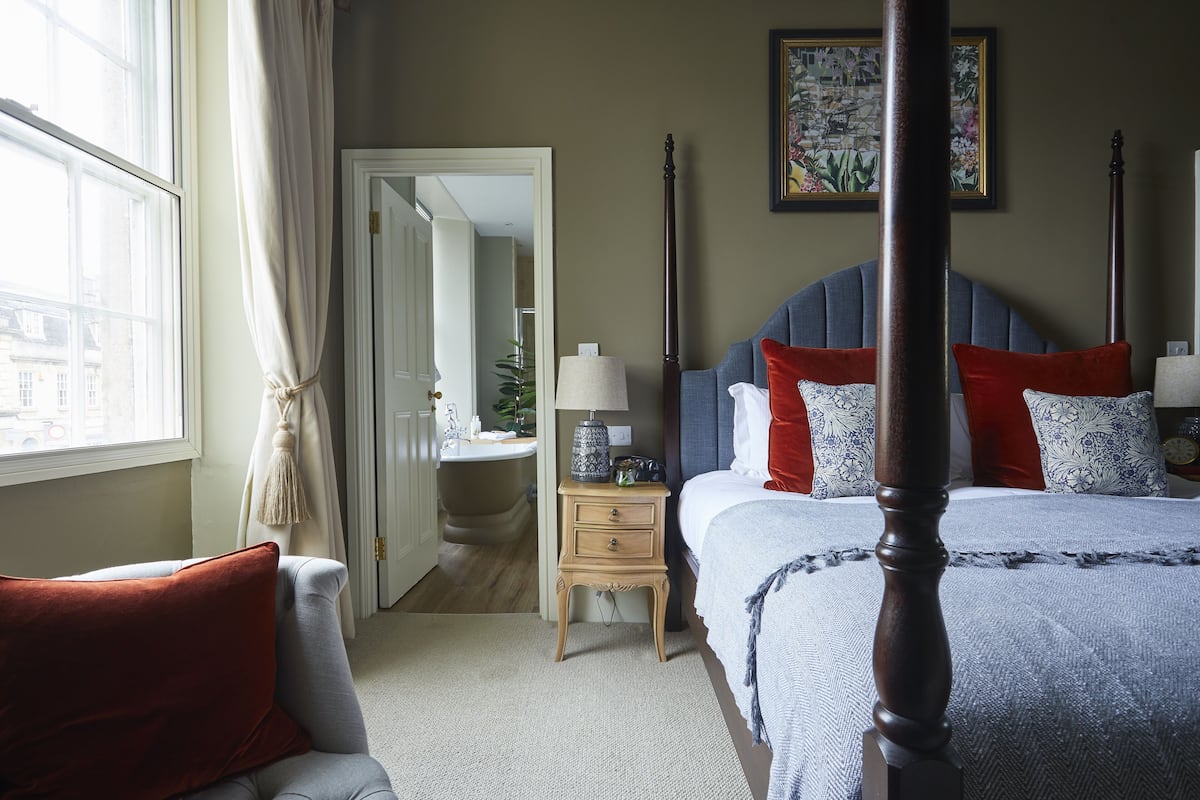 A bedroom with a four-poster bed, a nightstand with a lamp, a chair with a red cushion, and a view of the city through a window.