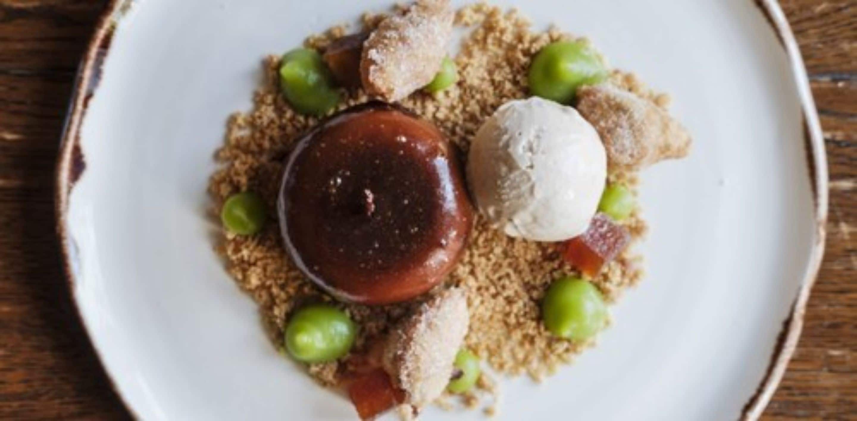 A dessert plate with a round chocolate dessert, a scoop of ice cream, and green grapes on a bed of crumbled biscuit.