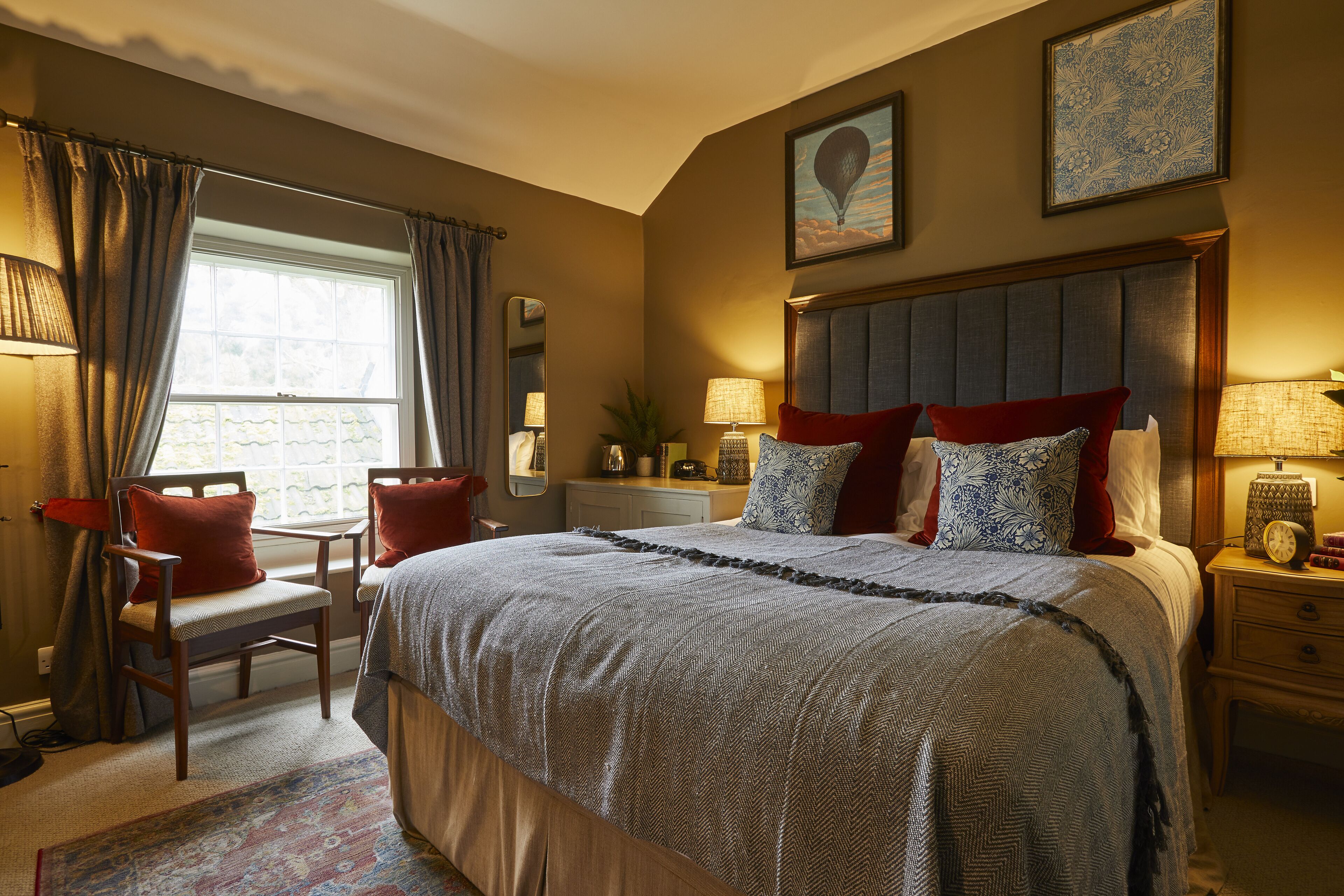 A well-lit bedroom with a large bed, a desk, and a chair. There is a window with curtains, a bedside table with a lamp, and a wall-mounted clock.