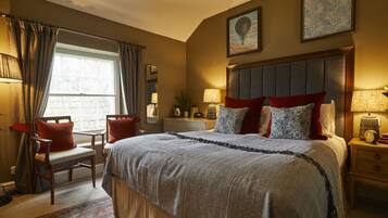 A well-lit bedroom with a large bed, a desk, and a chair. There is a window with curtains, a bedside table with a lamp, and a wall-mounted clock.