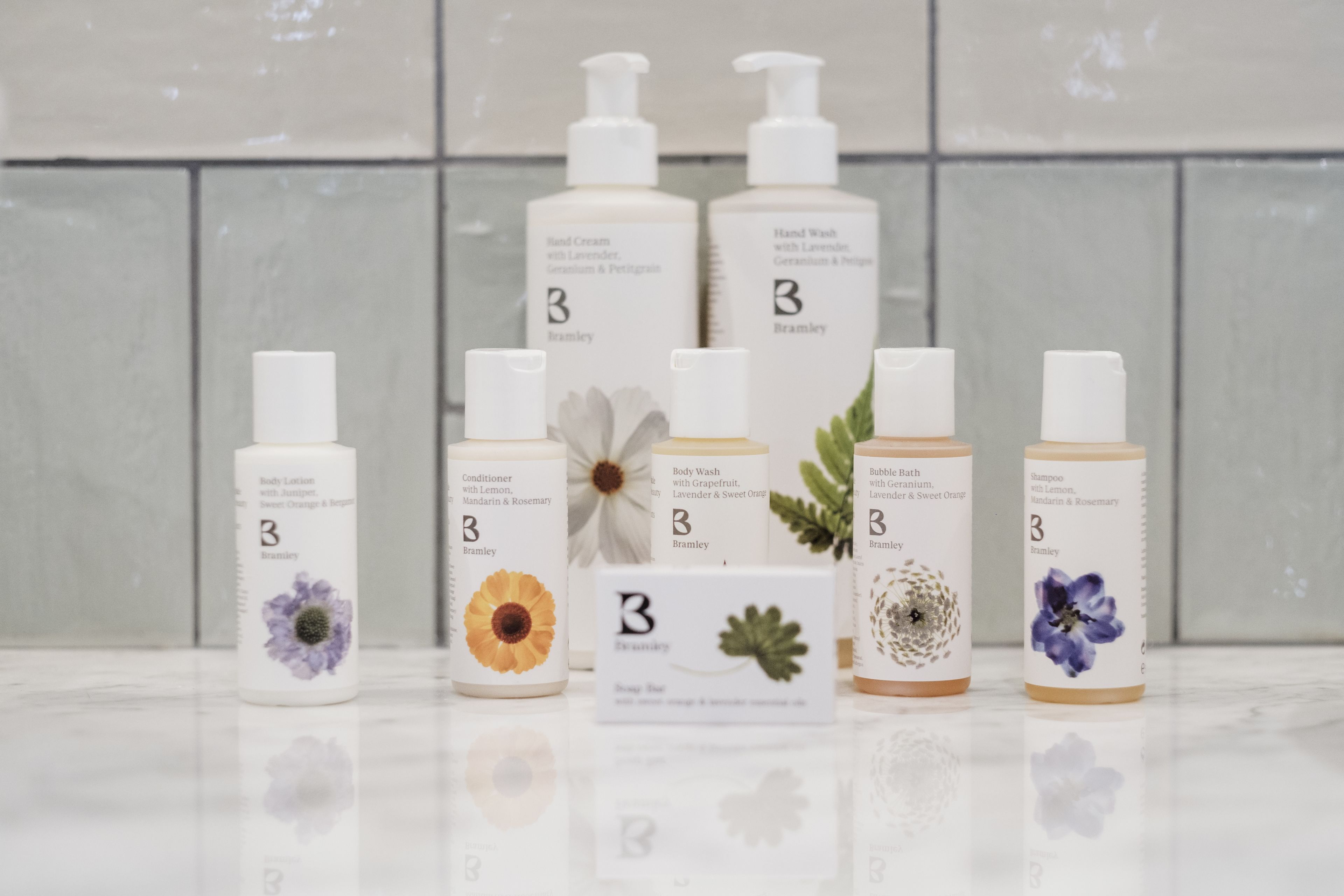 A selection of skincare products with floral designs on their labels, arranged on a reflective surface against a tiled wall background.
