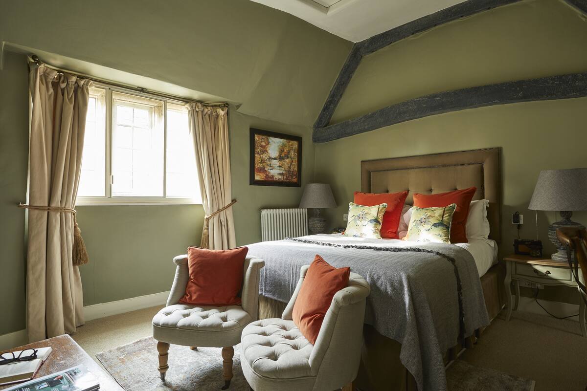 A bedroom with a bed, two chairs, a nightstand, a lamp, and a window with curtains.