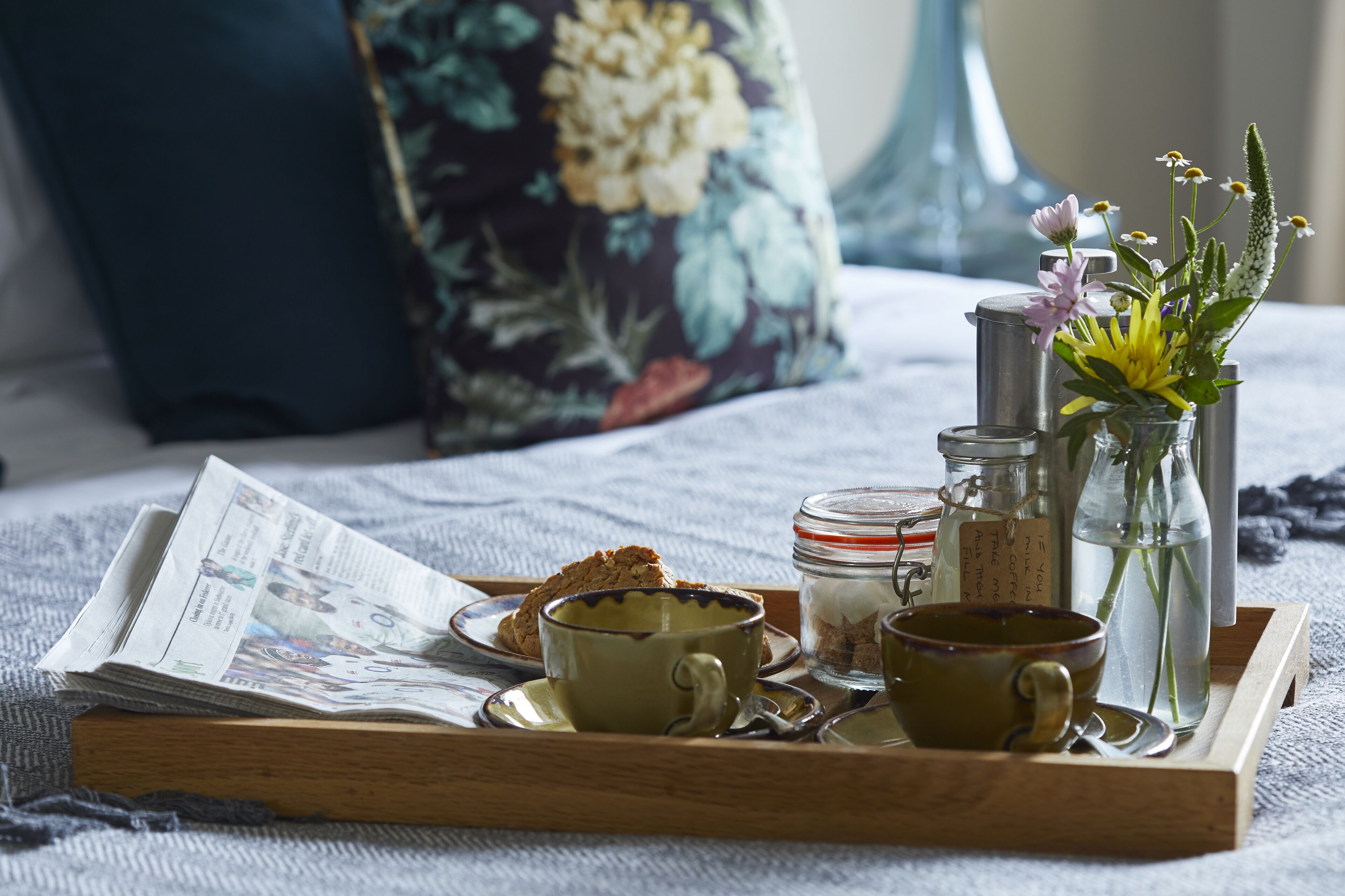 A breakfast tray with a newspaper, two cups, a jar, and a bottle on a bed with floral pillows.