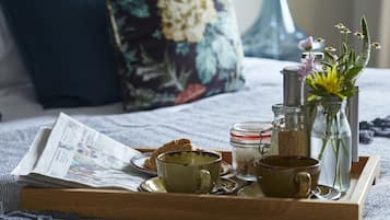 A breakfast tray with a newspaper, two cups, a jar, and a bottle on a bed with floral pillows.