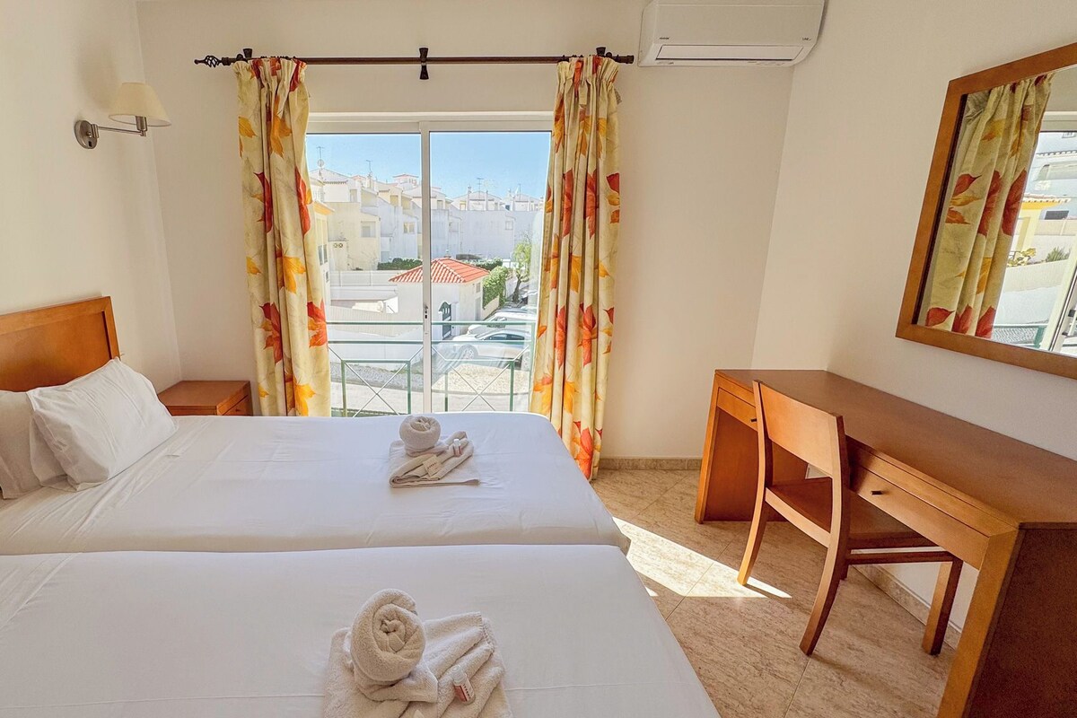 A hotel room with two beds, a desk, a chair, a window with curtains, and a painting on the wall.