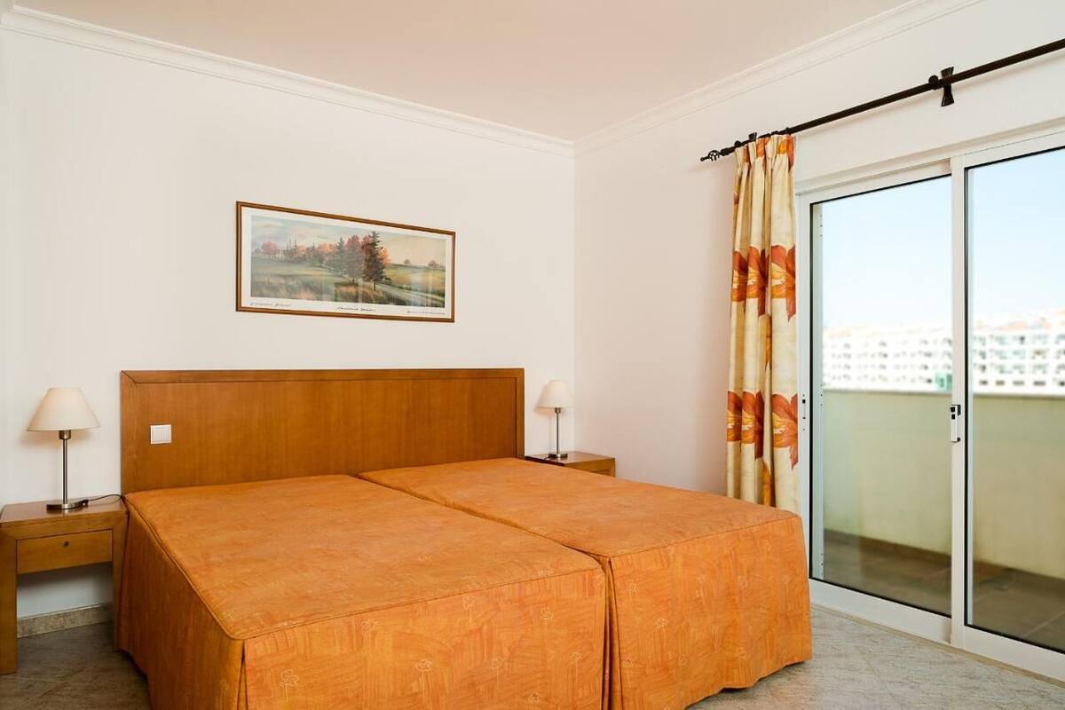 A double bed with a wooden headboard, two bedside tables with lamps, a framed landscape painting on the wall, and a sliding glass door to a balcony.