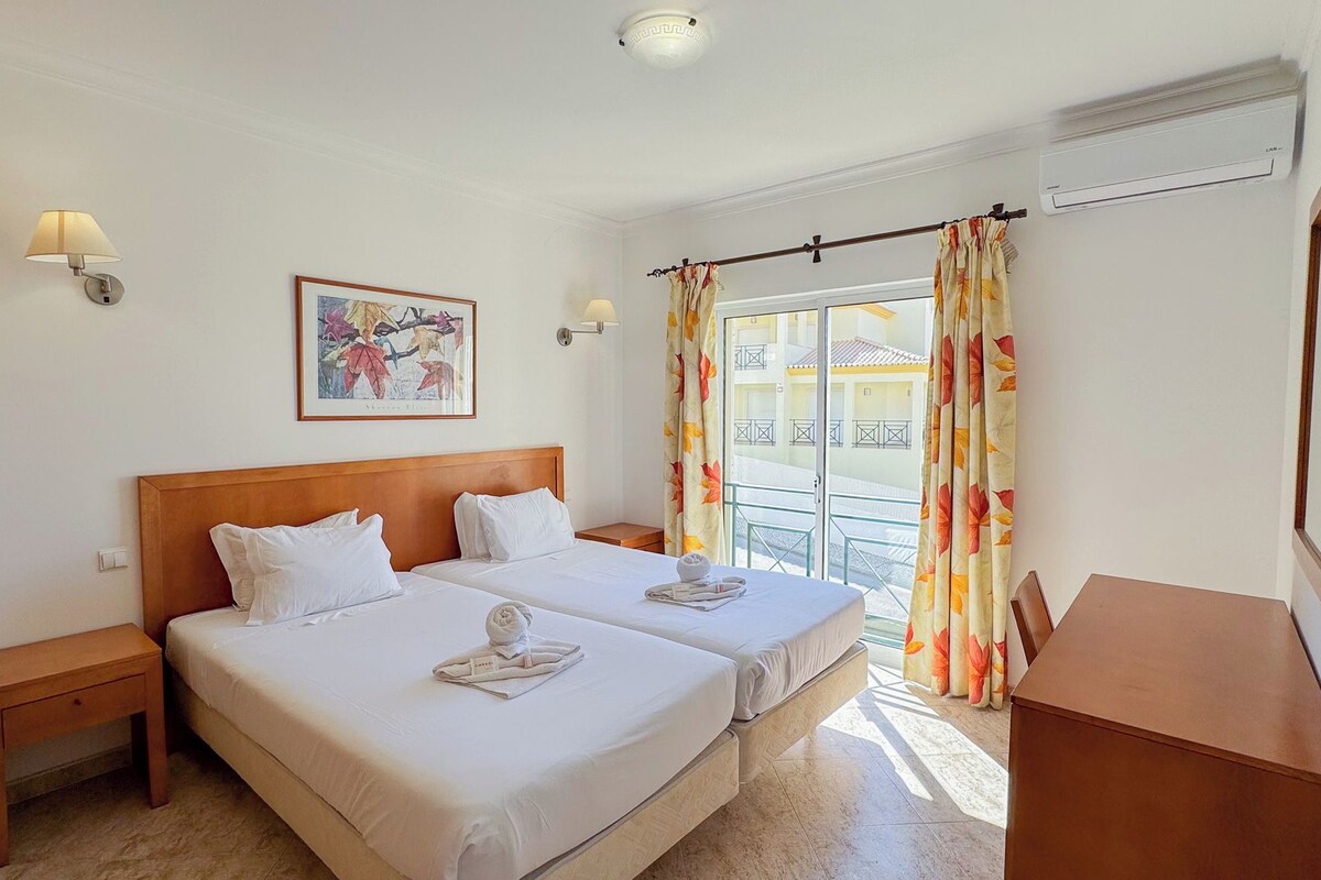 A hotel room with two beds, a wooden headboard, a painting, a window with curtains, and an air conditioning unit.